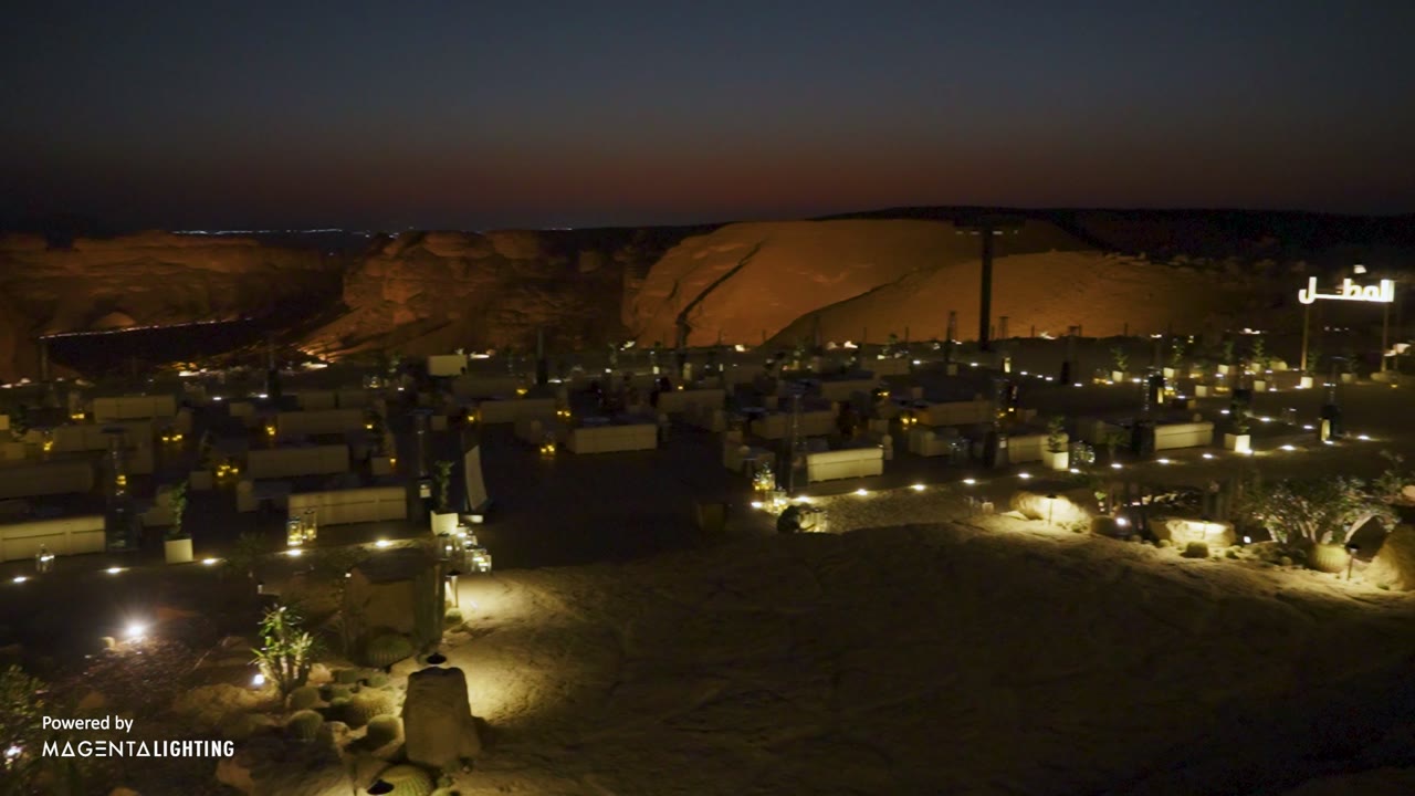 A desert rock face lit warmly from below at night, with a set dining terrace on the sand beneath it.