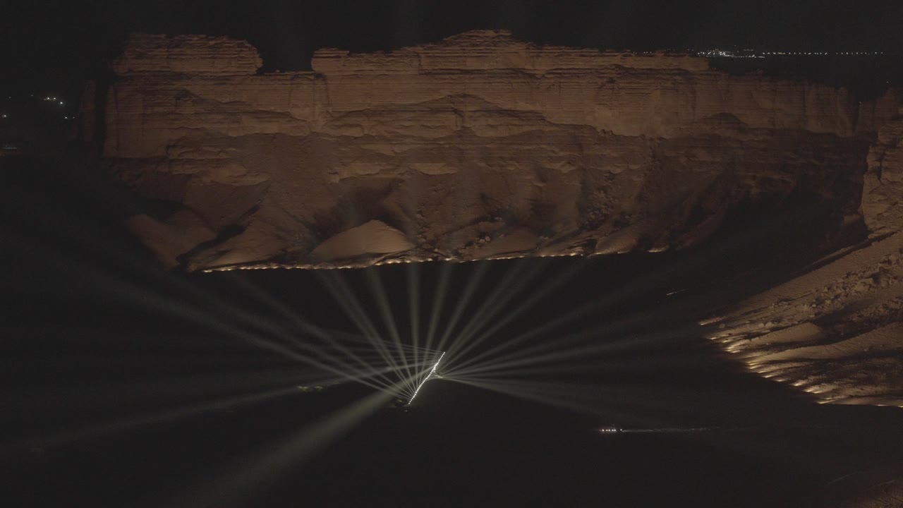 A fan of white beams opening out from a stage on the desert floor, beneath a floodlit rock escarpment at night.