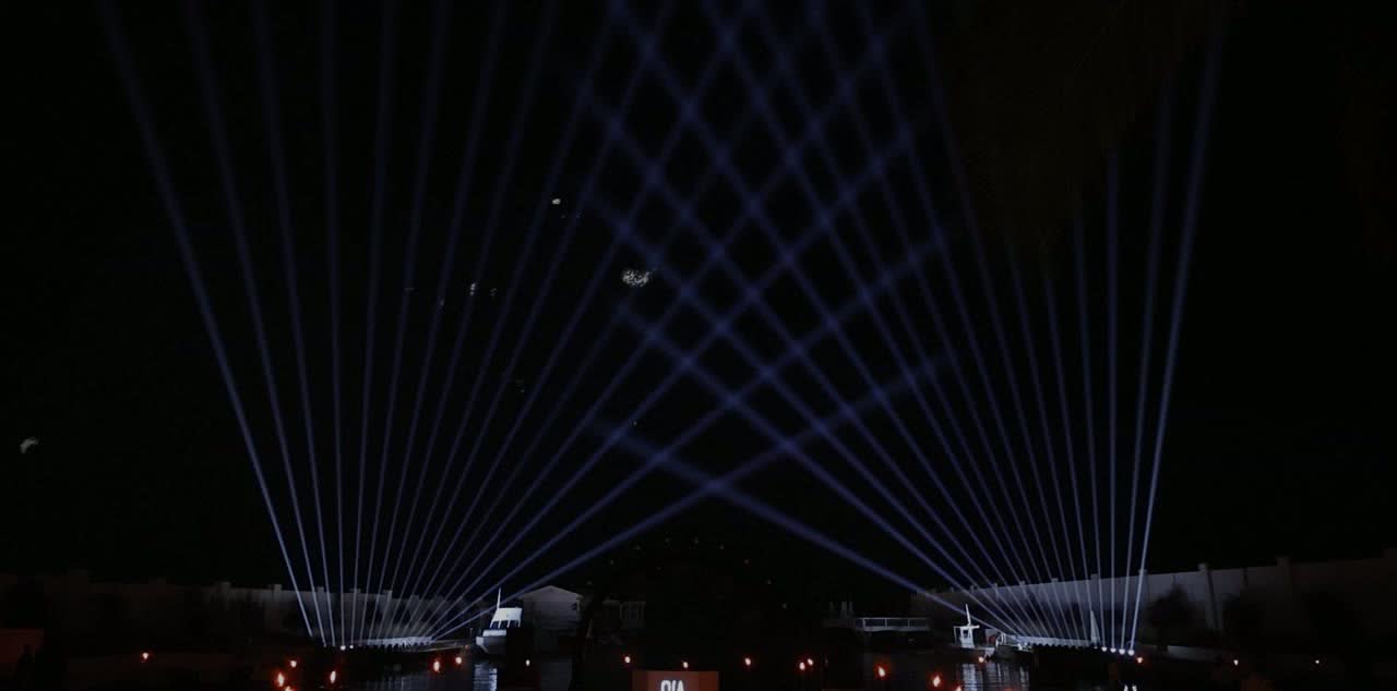 Blue-white beam fans opening above an open-air beach resort at night, reflected across the water in front of it.